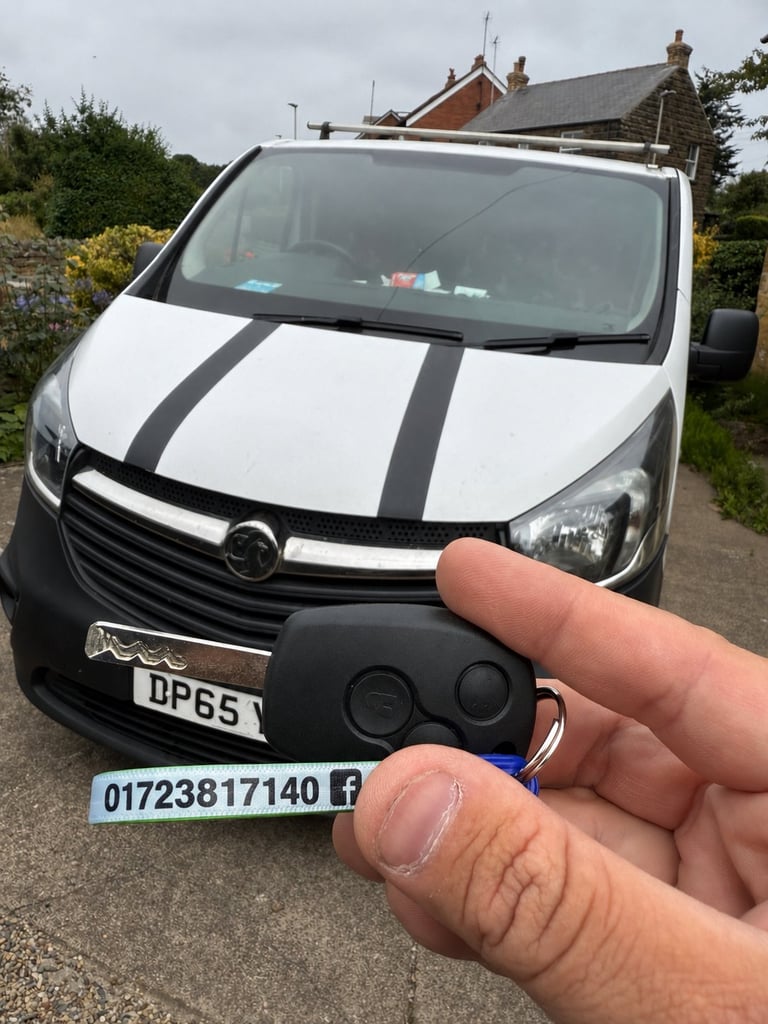 Hand holding a Vauxhall car key fob in front of a white Vauxhall Vivaro van with black racing stripes parked on a residential drive
