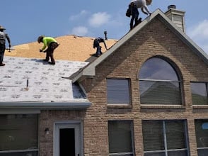 Workers in safety vests installing roofing materials on a brick house with a large arched window