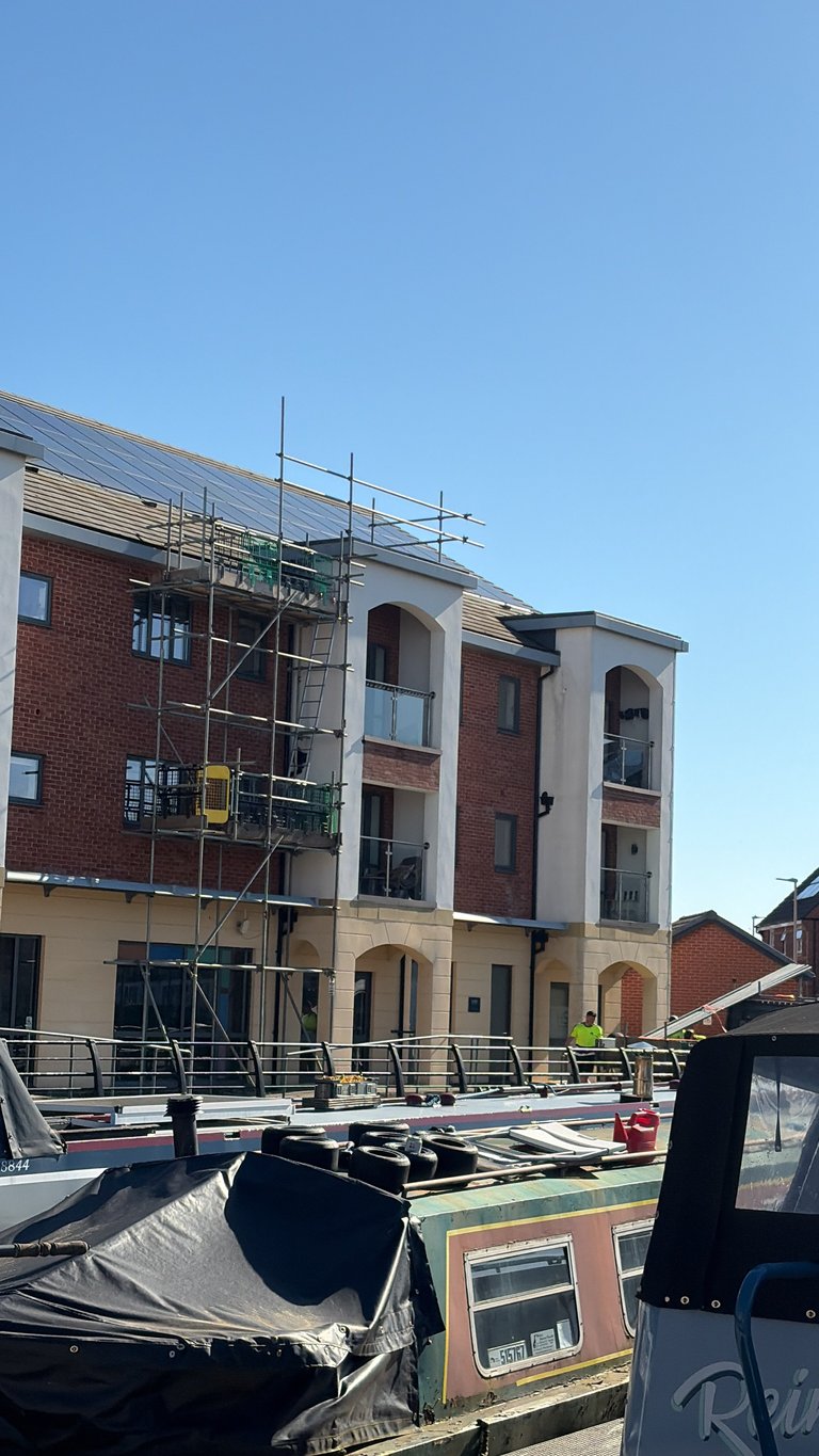Multi-story brick building under construction with metal scaffolding on roof and canal boats in foreground
