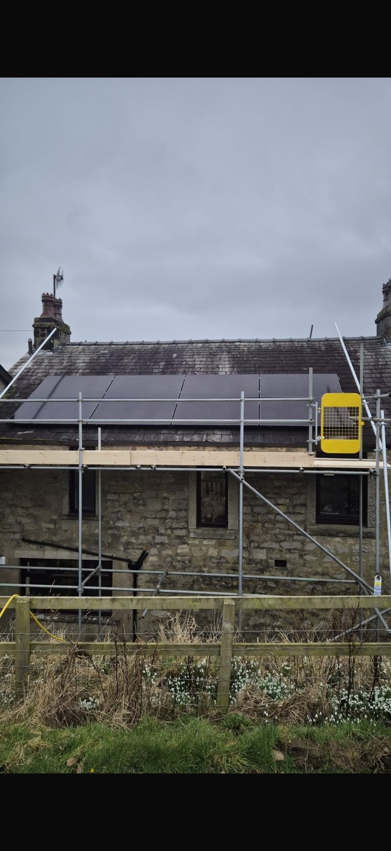 Stone cottage under renovation with solar panels on roof and metal scaffolding, enclosed by wooden fence against overcast sky