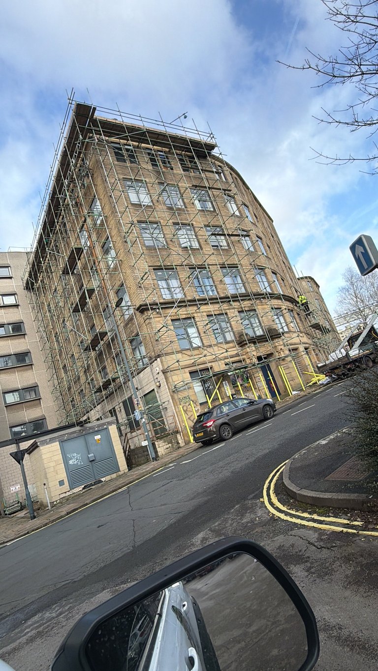 Multi-story brick building under construction with metal scaffolding on top, street view with parked car and bare trees