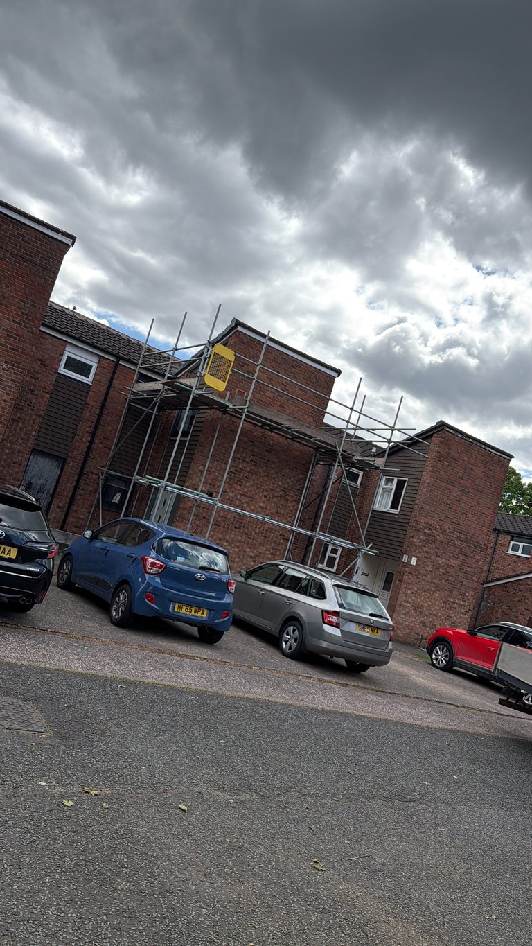 Brick residential building with scaffolding under renovation, parked cars in driveway, dramatic cloudy sky overhead