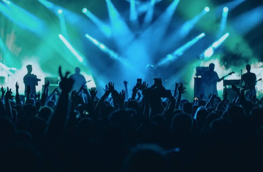 Silhouetted crowd with raised hands at a concert with blue stage lighting and band performers on stage