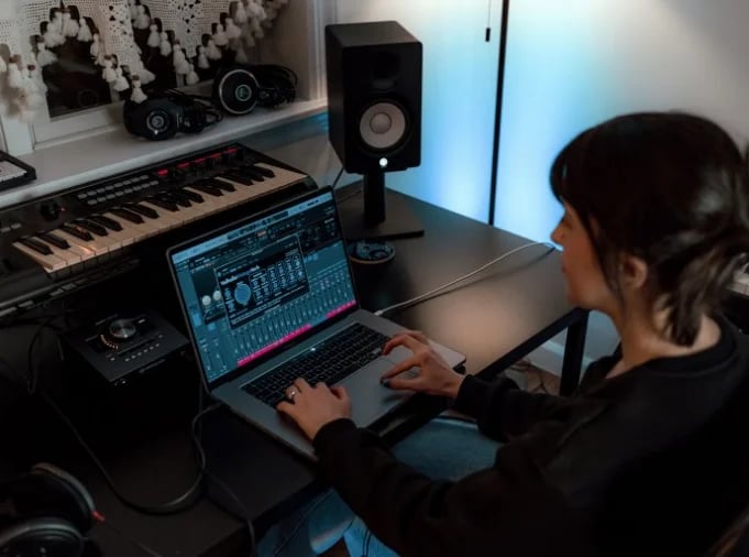 Person working at music production setup with laptop displaying audio software, keyboard, and studio monitor speaker