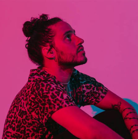 Man with long hair and beard wearing a leopard print jacket against a bright pink background, looking over his shoulder