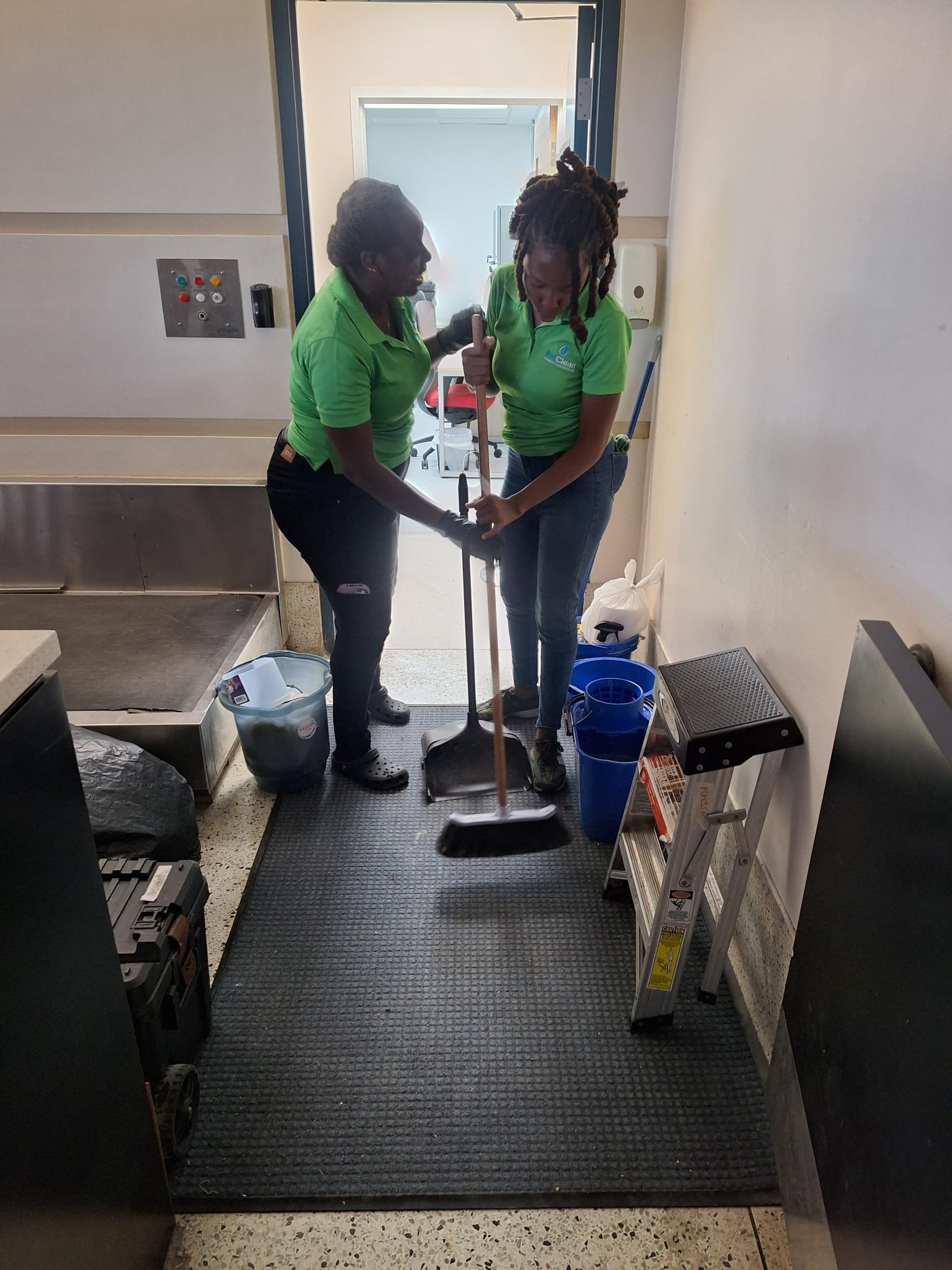 Two cleaning workers in green shirts sweeping a commercial kitchen floor with cleaning supplies visible nearby