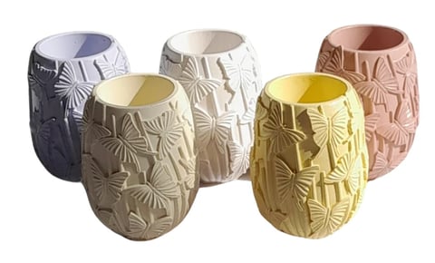 Five decorative candle vessels in different colors with embossed butterfly patterns and colored wax interiors arranged in a row