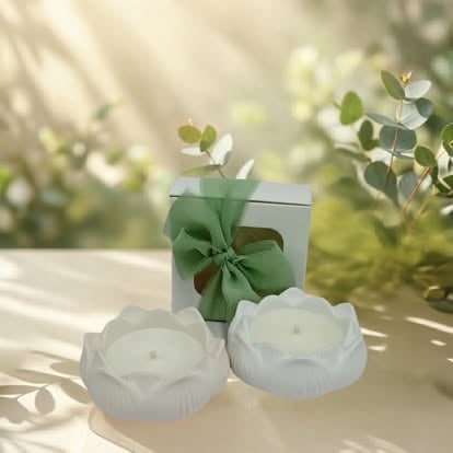 White gift box with green ribbon bow positioned behind two white frosted rose-shaped candles on a sunny surface with blurred green foliage background