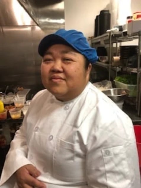 A man wearing a white chefs jacket and blue cap smiling in a commercial kitchen