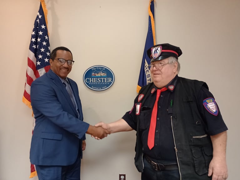 Two men shaking hands in front of American and Chester city flags in an official setting