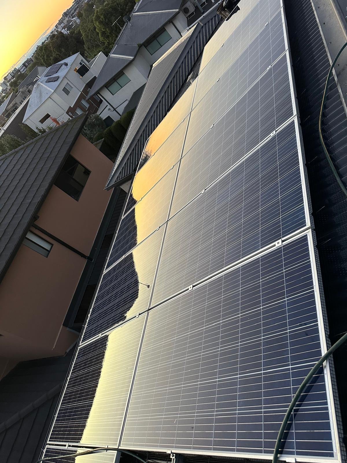 Large solar panel array mounted on residential rooftop with homes visible in background at sunset