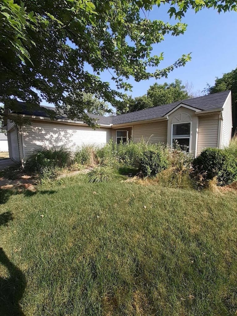 Single-story house with beige siding, attached garage, and overgrown lawn with trees overhead.