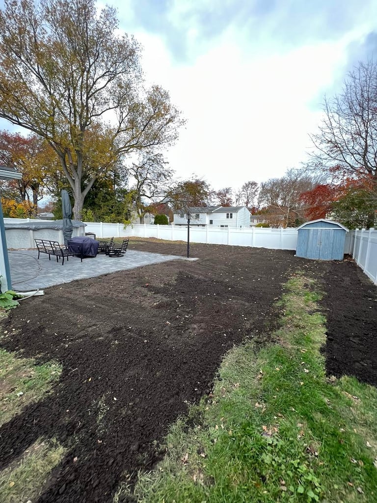 Backyard with freshly tilled dark soil garden bed, white fence, patio seating area, and bare autumn trees