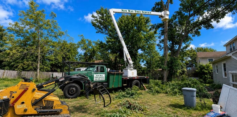 Green and white bucket truck with boom lift parked in residential yard next to yellow skid steer loader
