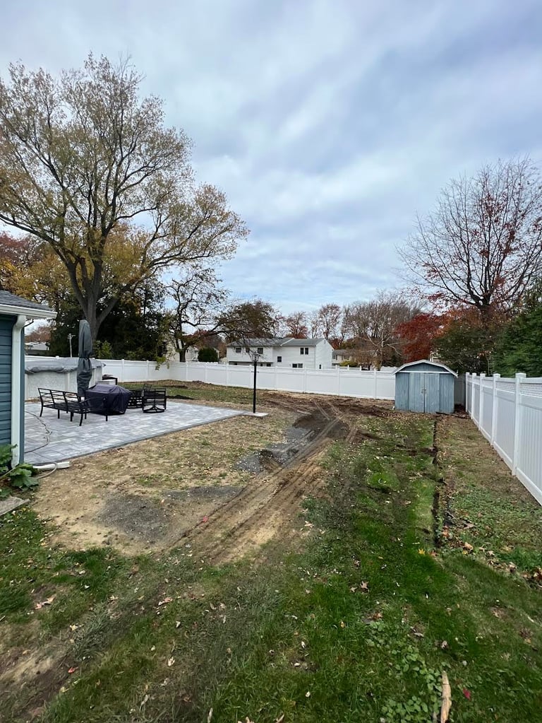 Suburban backyard with patio, bare trees, white fence, and distant houses under cloudy autumn sky