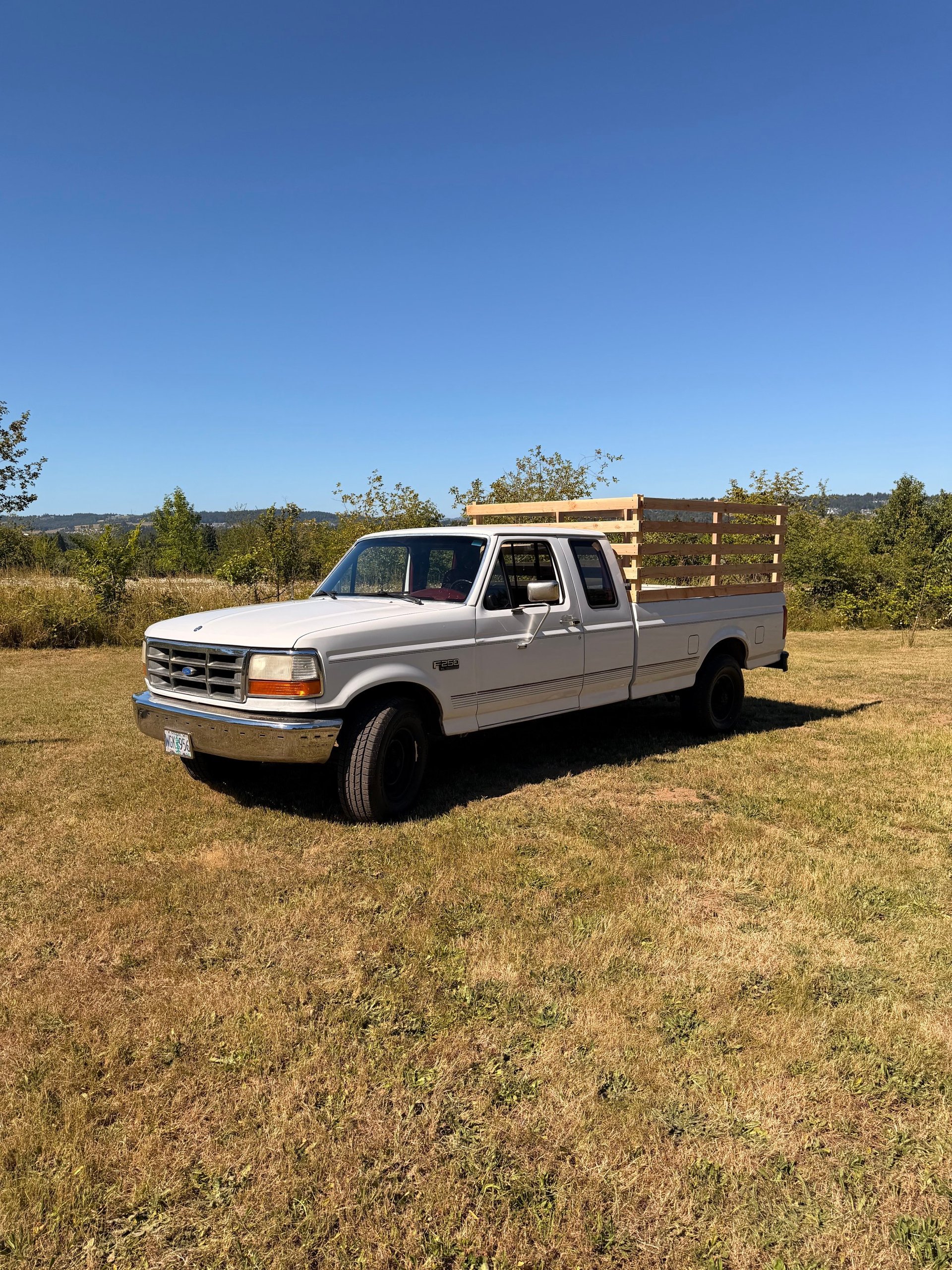 White Ford pickup truck with wooden stake bed parked in a grass field with trees and hills in the background