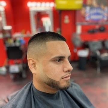 Man with short cropped hair and facial hair wearing a dark shirt, photographed in a barber shop with red walls in the background