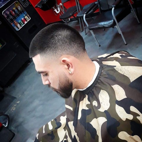 Man with short dark hair and beard wearing camouflage patterned shirt, photographed from above in an indoor barbershop setting