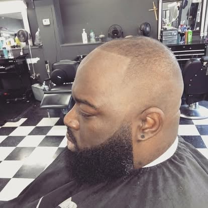 Man with shaved head and long beard in profile inside a barbershop with checkered floor