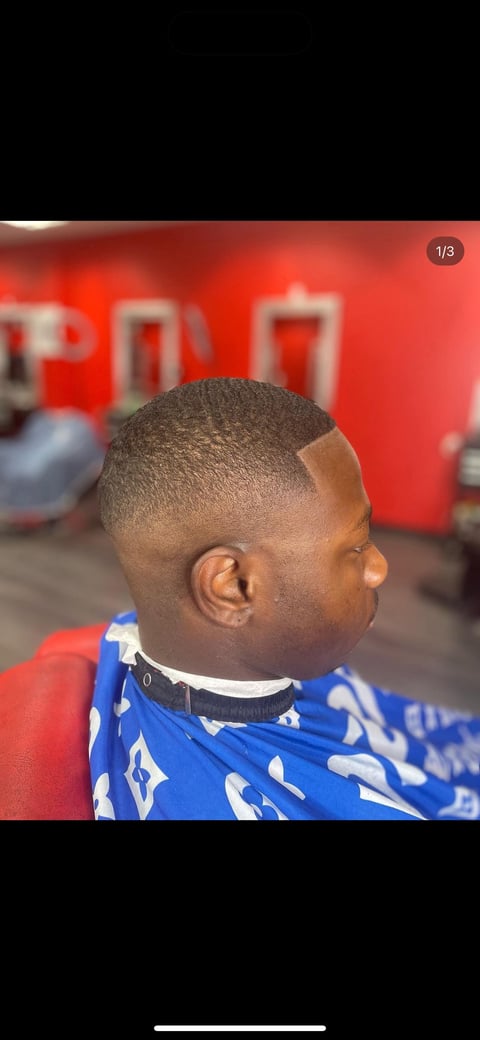Side profile of a person with a short, closely cropped haircut at a barbershop with red signage in the background
