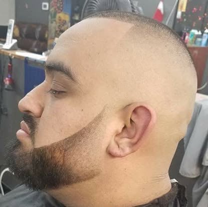 Side profile of a man with a closely shaved head, dark beard, and mustache in a barber shop setting