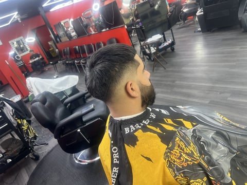 Man with beard in yellow and black shirt sitting in barber chair in salon with red furniture and equipment in background