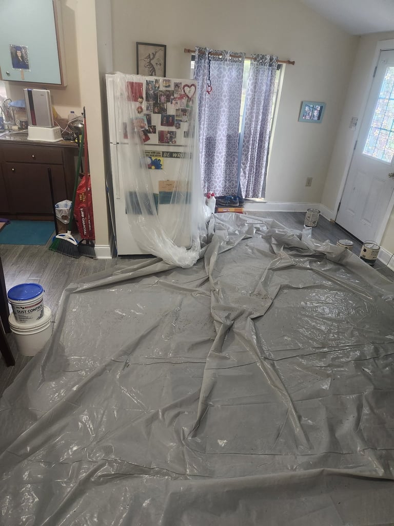 Interior room under construction with plastic sheeting covering the floor and furniture, paint supplies visible, and windows in background