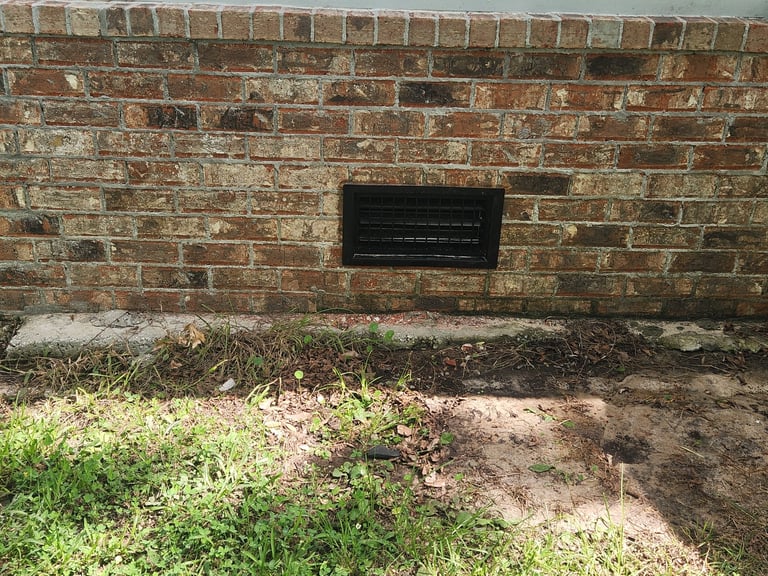 Weathered brick building foundation with black vent grate and overgrown grass and weeds below
