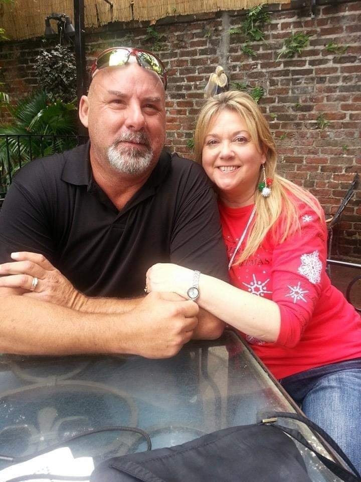 A man in a black polo shirt and a woman in a red sweater with snowflake patterns smile together outdoors against a brick wall