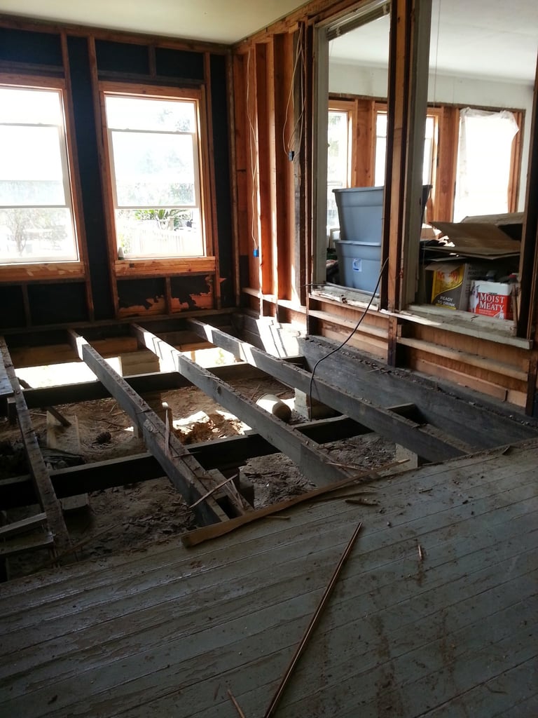 Interior of a home under renovation with exposed floor joists and wooden window frames with brown curtains