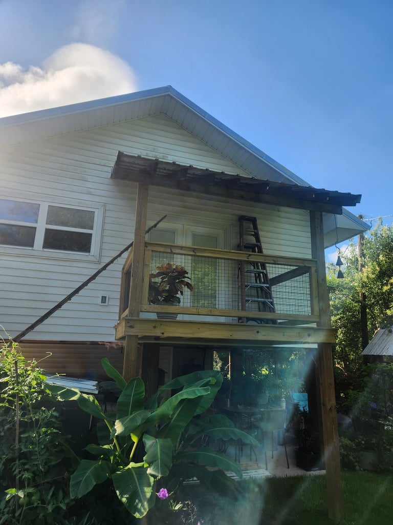 Elevated beach house with gray siding, wooden deck, and lush green vegetation below under clear blue sky
