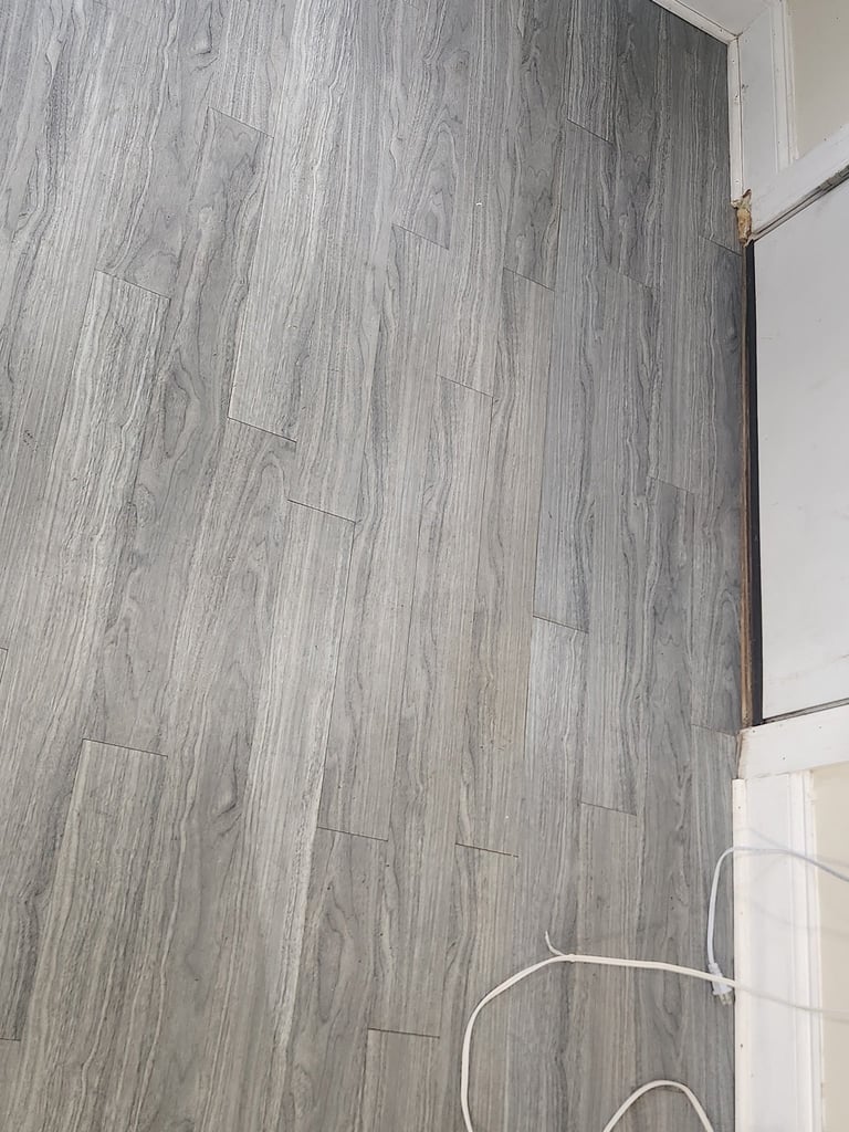 Gray wood-grain laminate flooring in a room with white trim and a white cable visible in corner