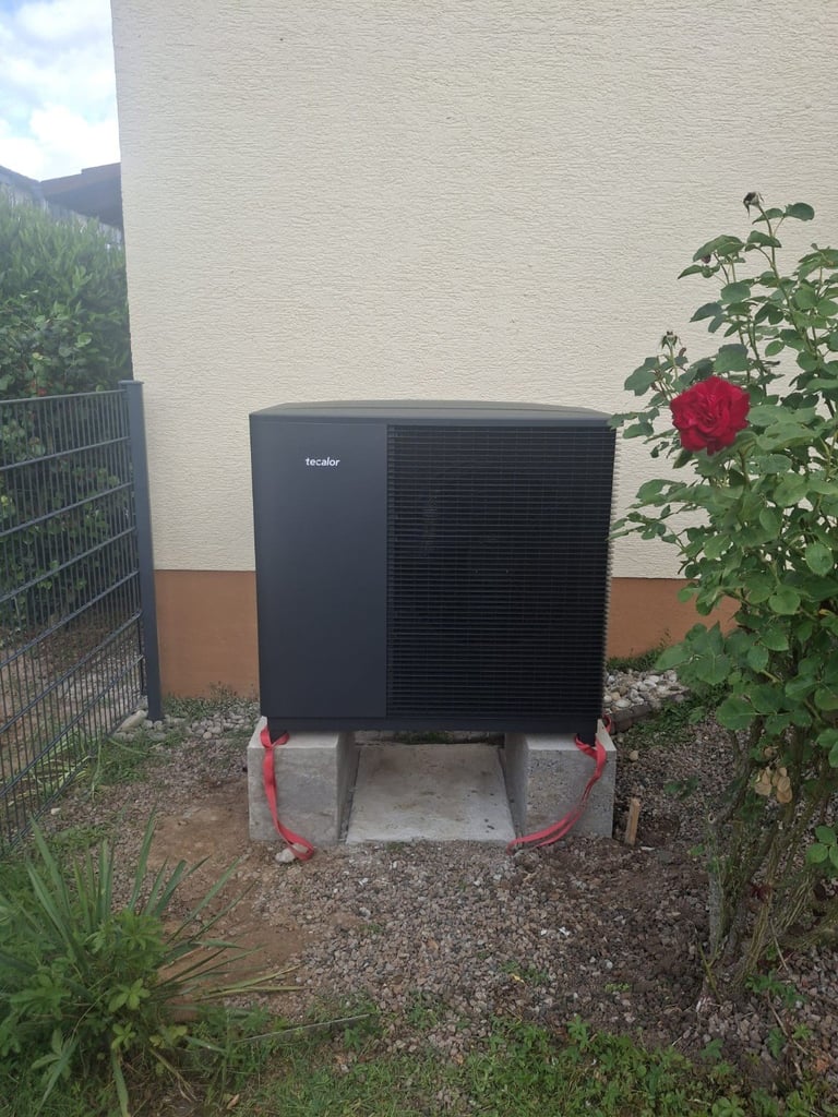 Black Hvelac heat pump unit installed outdoors against a cream and brown wall with red roses and garden surroundings