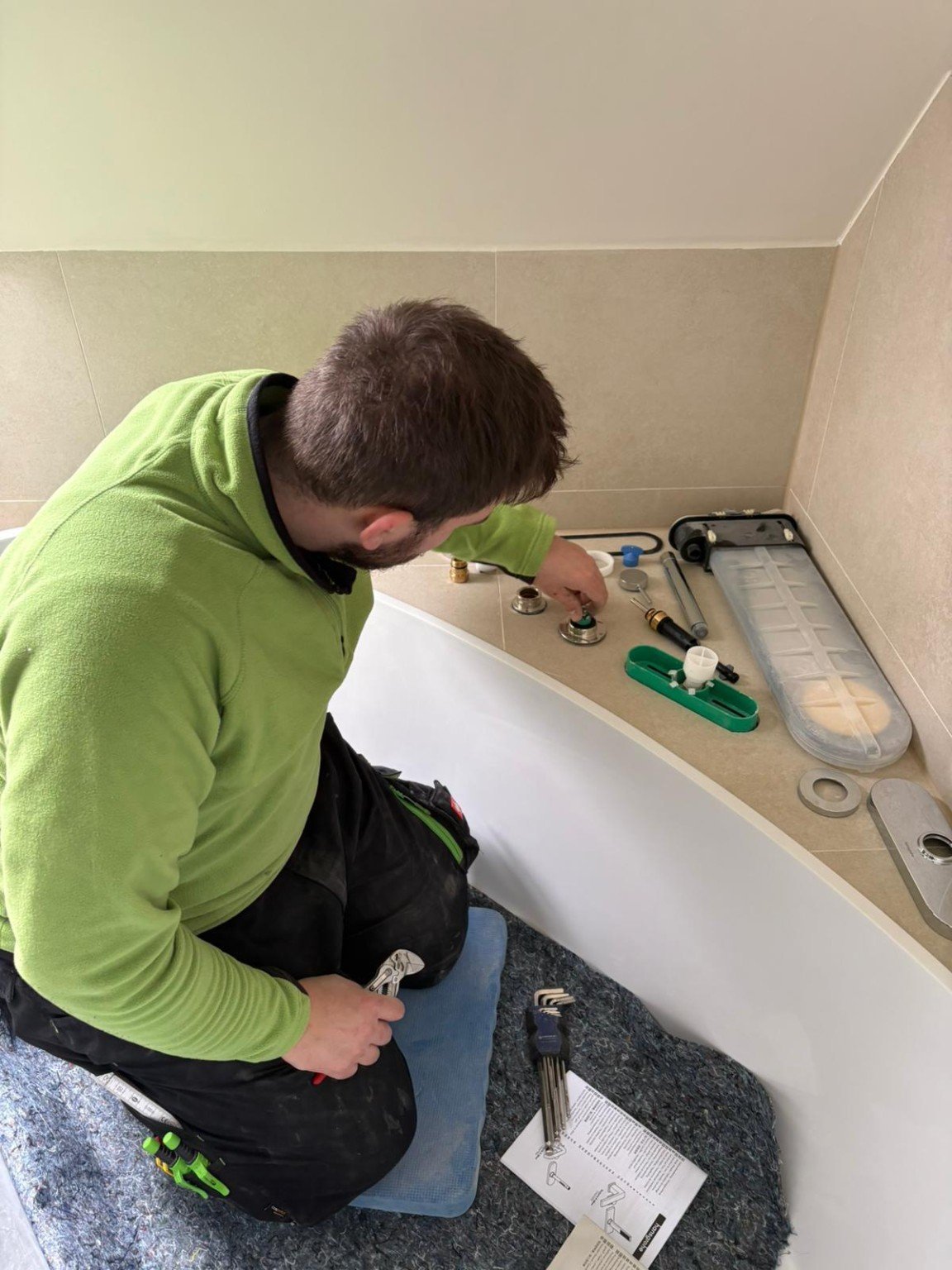 Technician in green jacket installing bathroom vanity faucet, with tools and supplies on counter