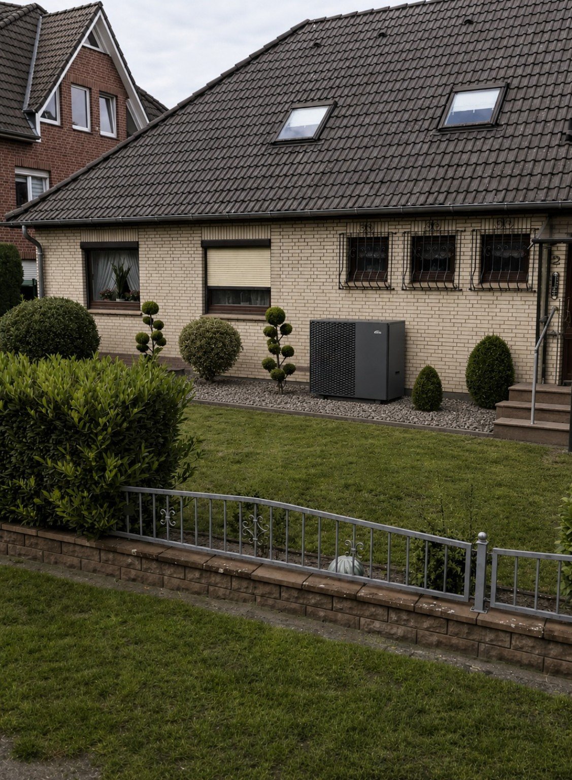 Modern brick house with dark tile roof, multiple windows, manicured garden with topiary, metal fence, and lawn in foreground