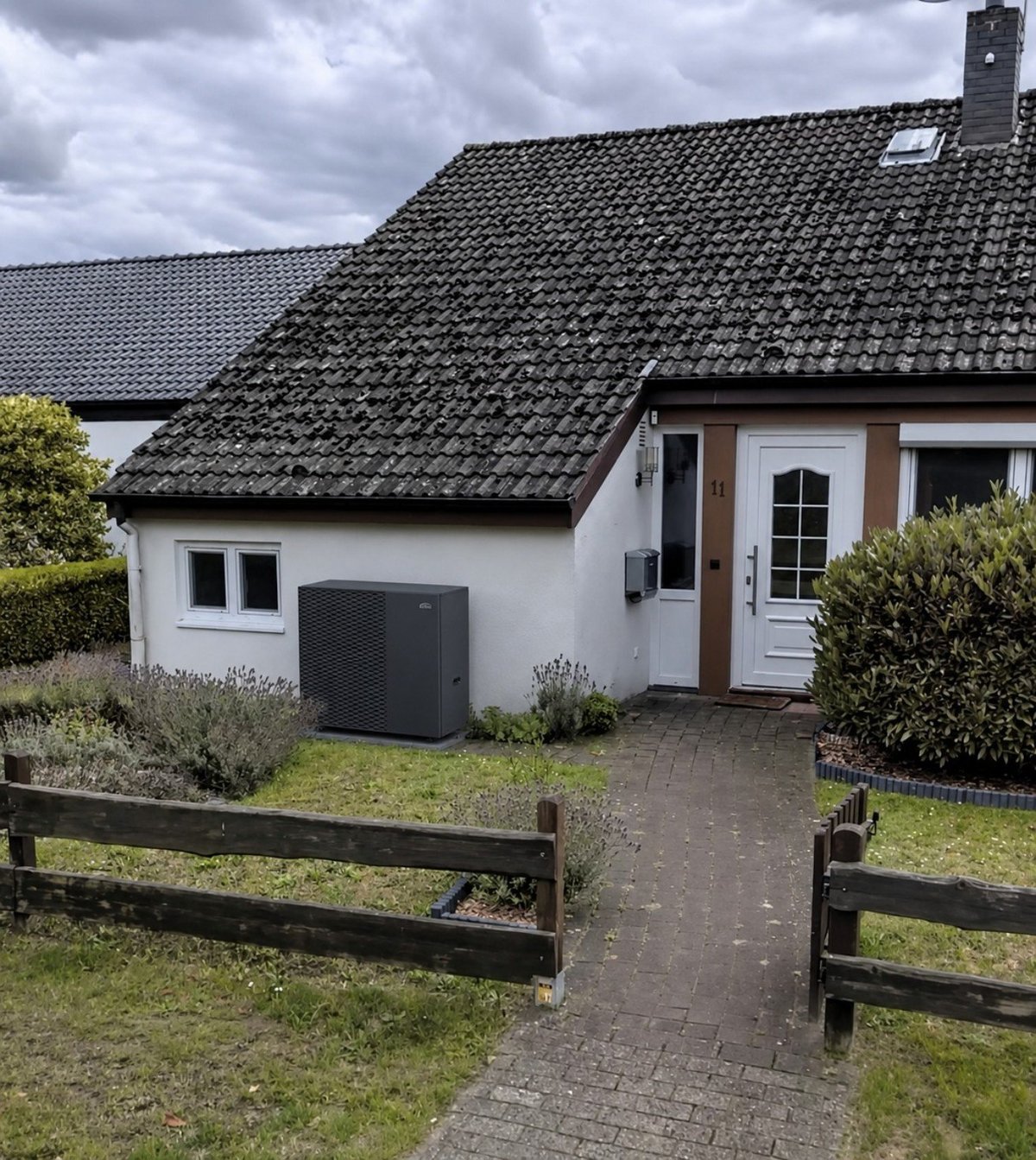 White cottage with dark tiled roof, wooden entrance door, surrounded by green hedges and wooden fence under cloudy sky