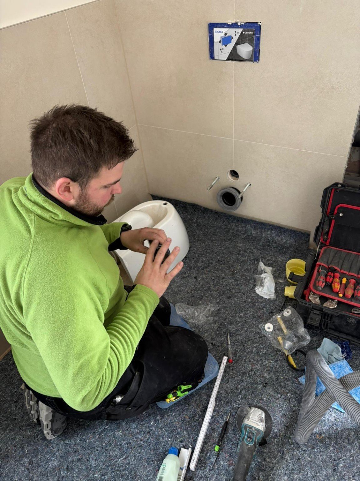Plumber in green shirt working on bathroom plumbing fixture, crouched with tools and toolbox nearby