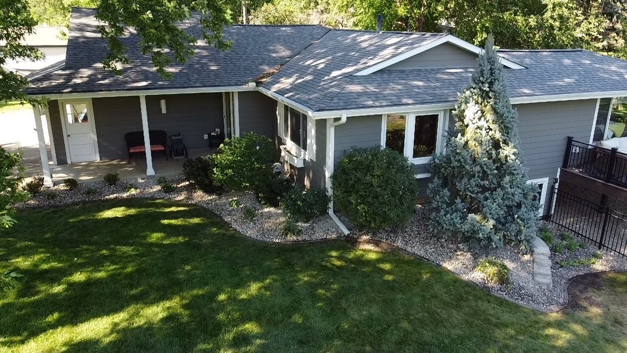 Aerial view of a single-story gray house with black roof, front porch, manicured lawn, landscaping, and driveway