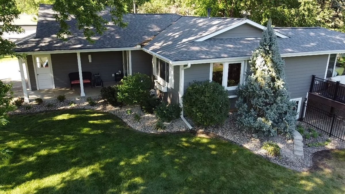 Aerial view of a single-story gray house with dark roof, covered porch, manicured lawn, and surrounding greenery and landscaping