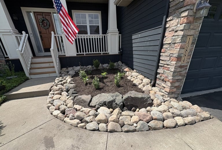 Front porch with boulder wall retaining dark mulch, plants, and river rock — after landscape redesign