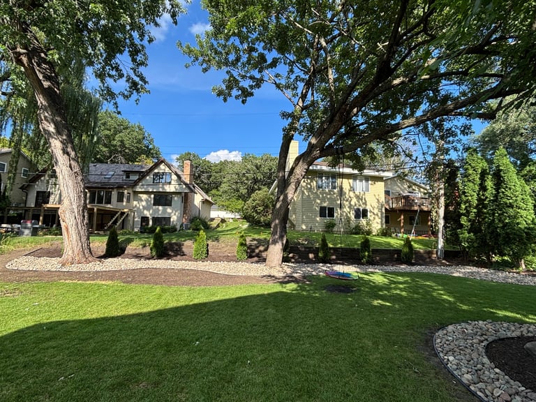 Large manicured lawn with stone pathways and mature trees, overlooking historic stone cottage buildings on a hillside under clear blue sky