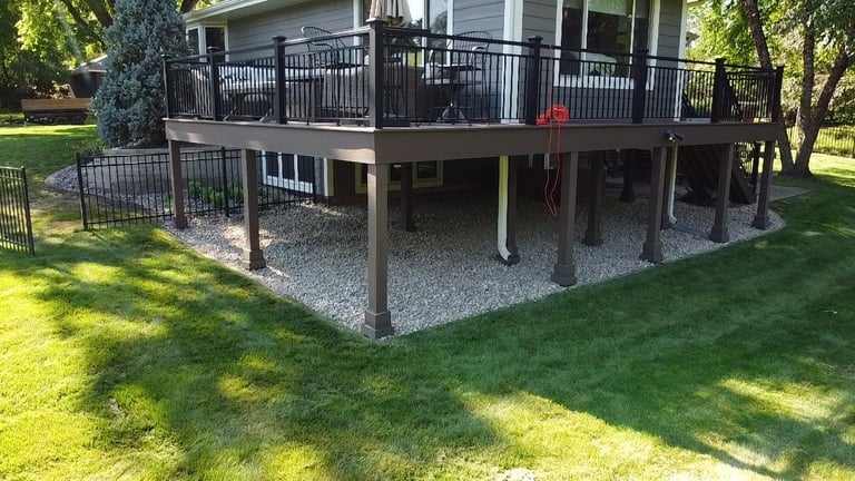 Two-story house with black deck railing and covered patio area with gravel underneath, surrounded by green lawn and trees — after renovation