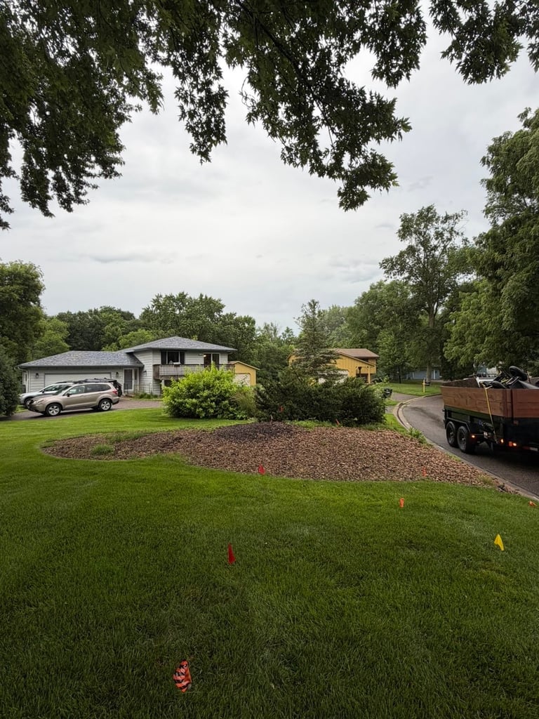 Residential home with driveway, parked car, and landscaped yard surrounded by mature trees on overcast day — before front bed work