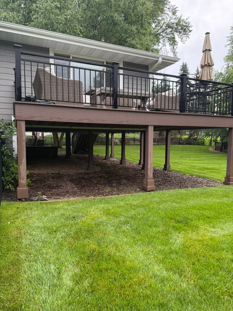 Modern elevated deck with black railings over a shaded patio area, attached to a residential home with green lawn and trees — before renovation