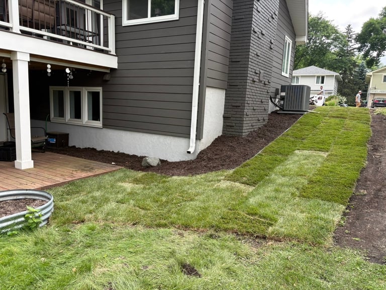 Backyard of a dark gray and white house with a wooden deck, green lawn, mulched garden beds, and neighboring homes visible in the background