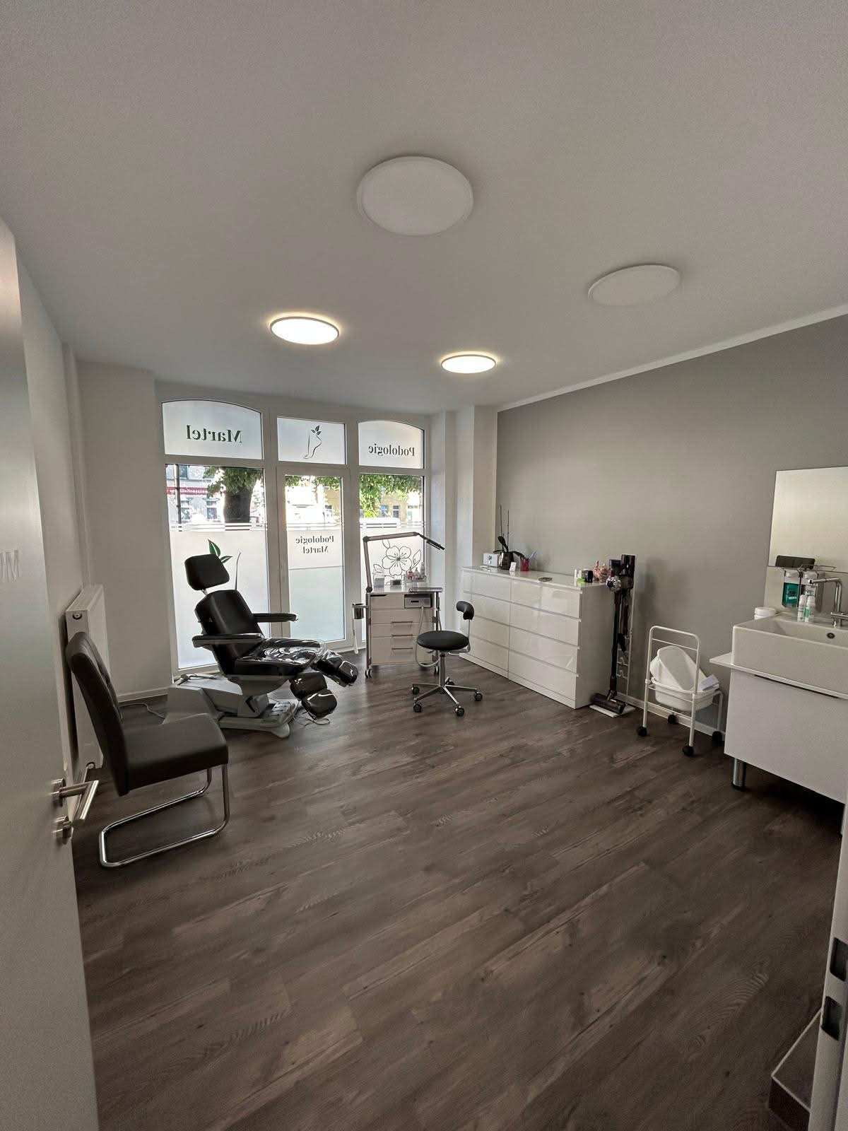 Behandlungsraum mit Podologiestuhl, Waschbecken und bodentiefen Fenstern