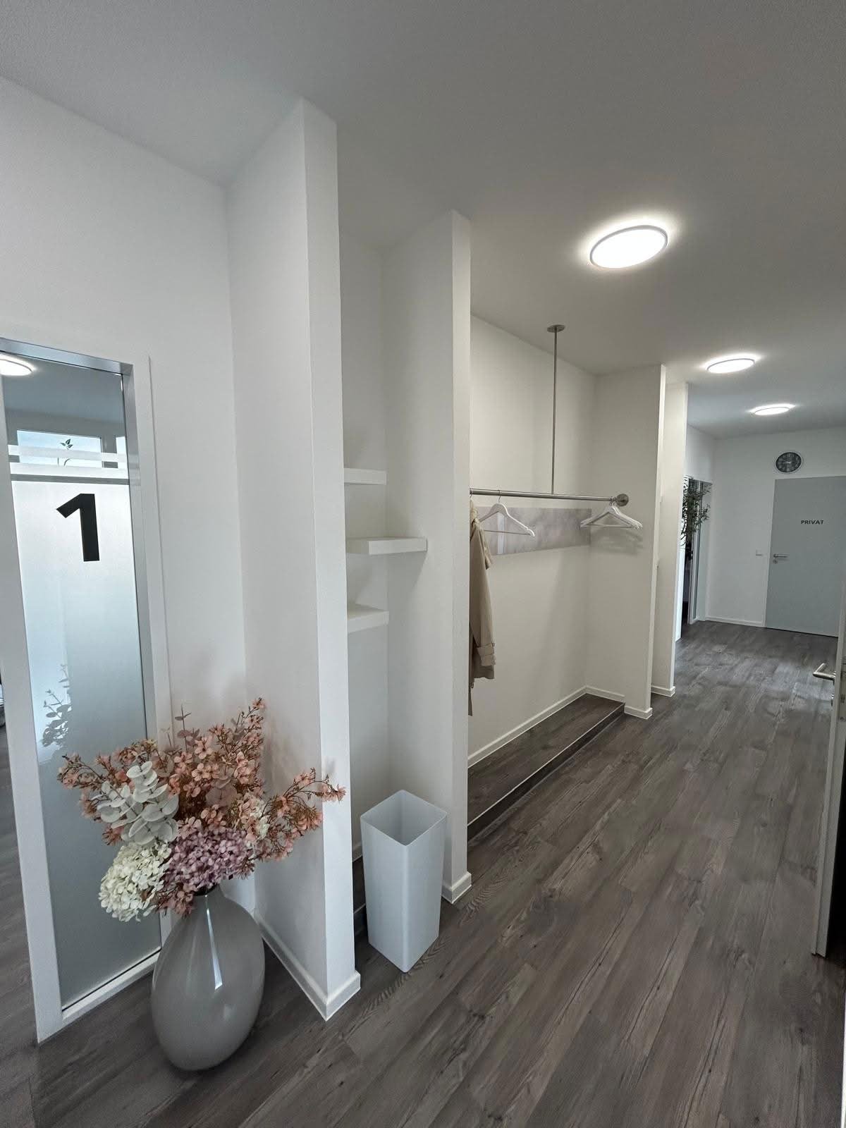 Modern hallway with white partitions, wooden flooring, dried flowers in a vase, and recessed ceiling lights