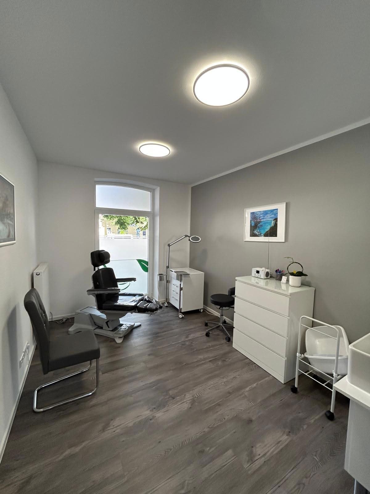 Modern medical office with dark wood flooring, white furniture, ceiling lights, and a door with natural light visible
