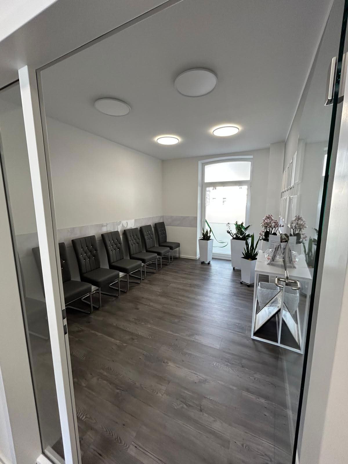 Modern medical waiting room with black chairs along walls, wood flooring, recessed lighting, and a window with potted plants on side table