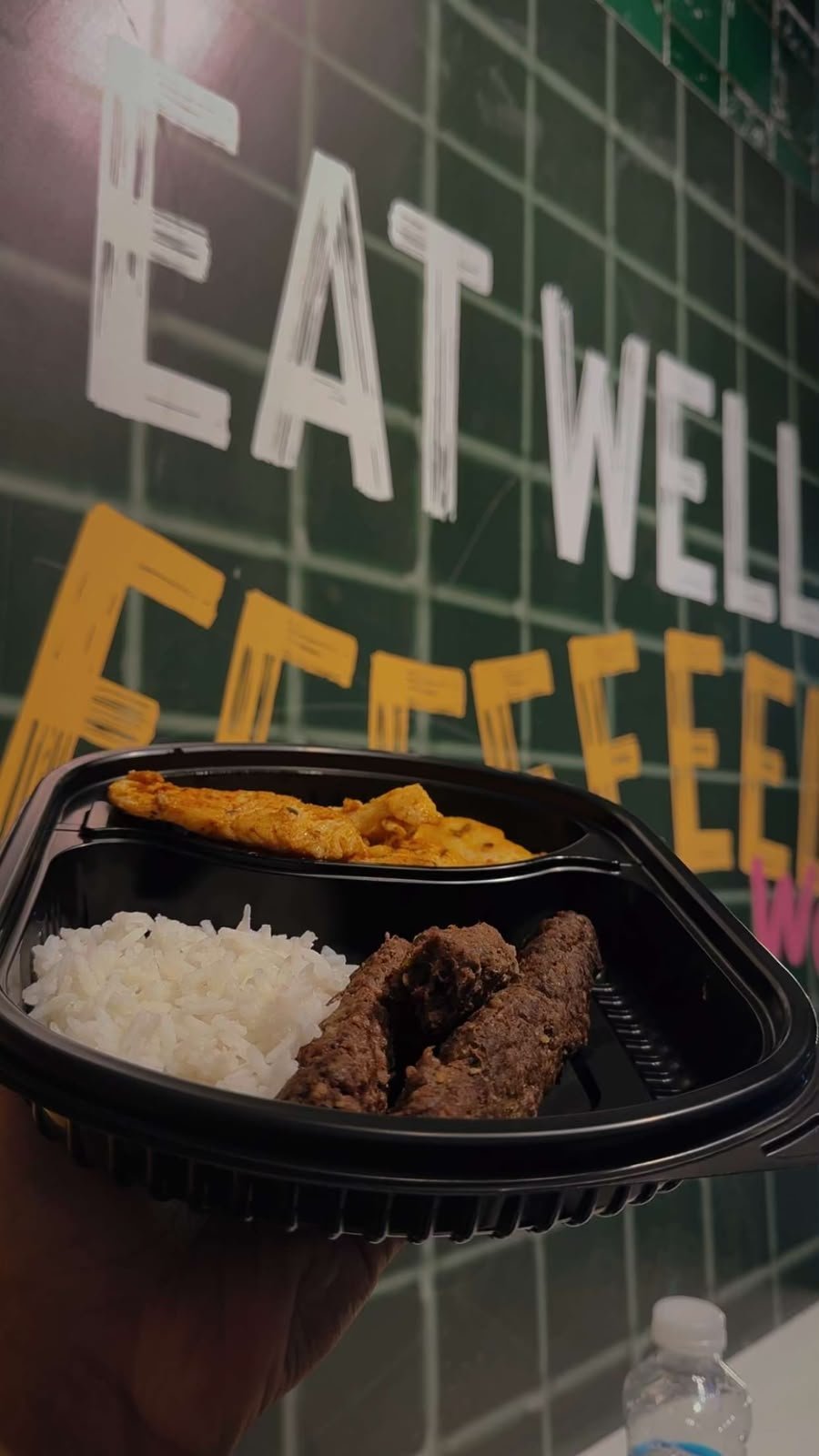 Black compartment tray with fried chicken, white rice, and plantains in front of a green wall with Eat Well signage